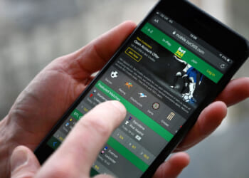 A person uses a smartphone to access a sports betting app, showing various sports icons and betting options on the screen.