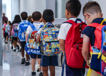 A group of young children, wearing backpacks and school uniforms, walk in a line down a brightly lit school hallway. The backpacks have colorful cartoon designs.