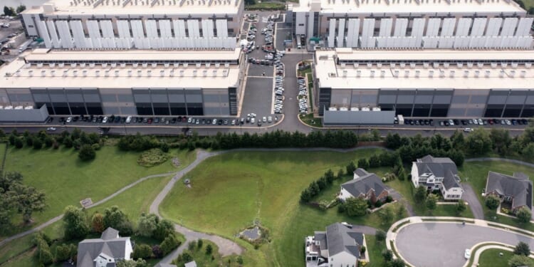 Aerial view of a data center with many parked cars, situated behind a green landscape with trees and several suburban houses at the bottom of the image.
