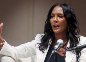 A woman with long dark hair and a white blazer speaks passionately into a microphone, gesturing with one hand while seated on a light-colored chair.