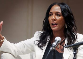 Gubernatorial candidate Keisha Lance Bottoms appears with long black hair, wearing a white blazer and gold hoop earrings. She speaks while gesturing with her hand in front of two microphones.