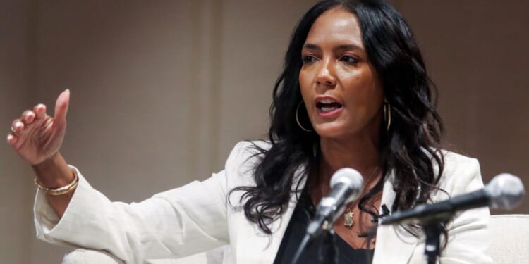 Gubernatorial candidate Keisha Lance Bottoms appears with long black hair, wearing a white blazer and gold hoop earrings. She speaks while gesturing with her hand in front of two microphones.