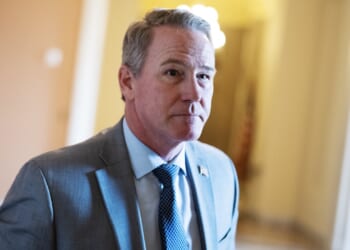 A man in a light gray suit and blue tie stands indoors, looking slightly to the side. The background is softly lit with warm colors and blurred details.