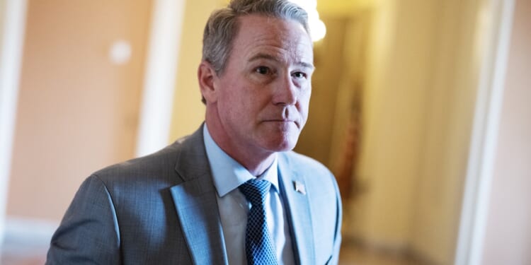 A man in a light gray suit and blue tie stands indoors, looking slightly to the side. The background is softly lit with warm colors and blurred details.
