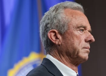 A man with gray hair in a suit stands in profile against a blue background with a blurred emblem or flag.