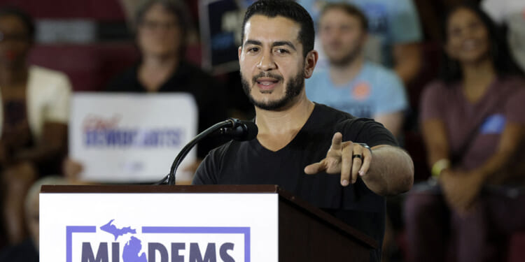 Michigan Democrat Abdul El-Sayed compared circumcision to transgender surgery. Pictured: El-Sayed, a man speaks at a podium labeled MIDEMS in front of an audience. He gestures with one hand while addressing the crowd. People in the background hold signs and appear to be listening attentively.