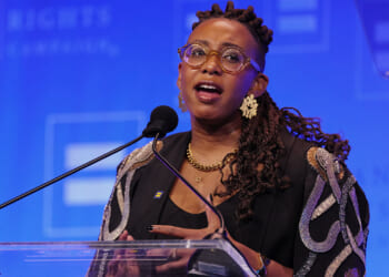The Human Rights Campaign condemned an HHS study finding that support for transgender medical procedures correlates with justifications for political violence. Pictured: HRC President Kelley Robinson, a person with glasses, braids, and gold jewelry speaks into a microphone at a podium, with a blue background featuring blurred text and logos.