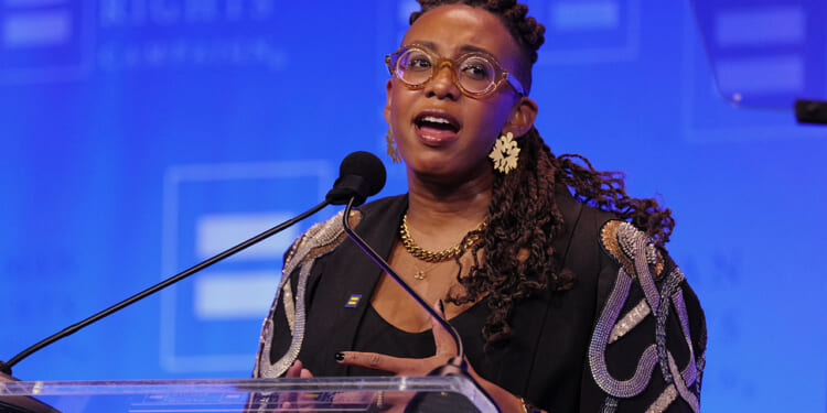 The Human Rights Campaign condemned an HHS study finding that support for transgender medical procedures correlates with justifications for political violence. Pictured: HRC President Kelley Robinson, a person with glasses, braids, and gold jewelry speaks into a microphone at a podium, with a blue background featuring blurred text and logos.