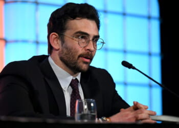 Hasan Piker in a suit and glasses speaks into a microphone at a panel, with his hands clasped on the table. Blue and orange backgrounds are behind him.