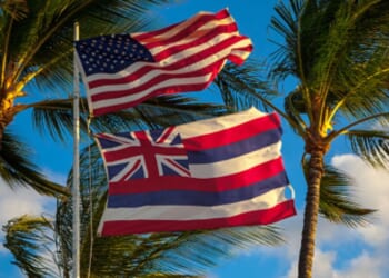 The American flag flies above the flag of Hawaii ahead of palm trees.