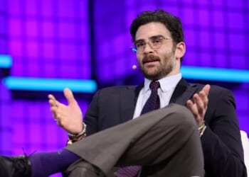 A man in a suit and tie, wearing glasses and a headset mic, gestures with his hands while speaking on stage against a purple backdrop with blue horizontal lights.