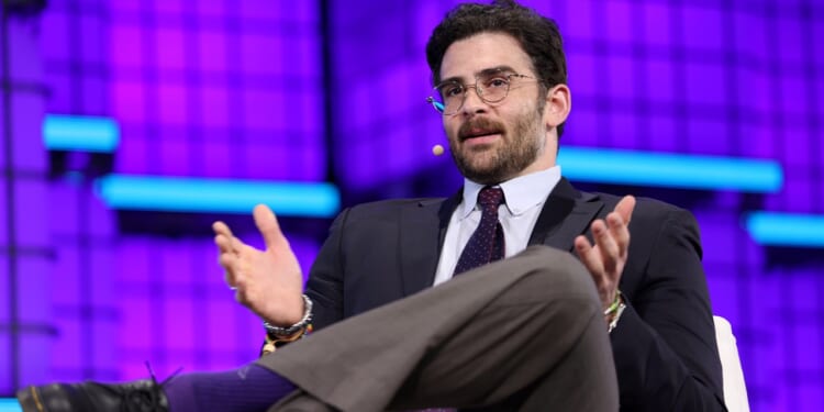 A man in a suit and tie, wearing glasses and a headset mic, gestures with his hands while speaking on stage against a purple backdrop with blue horizontal lights.