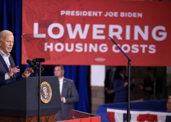 President Joe Biden speaks at a podium with a red sign behind him that reads Lowering Housing Costs during an event.