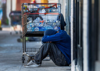 A person in worn clothes sits on a sidewalk with their head on their knees, next to a shopping cart filled with bottles and bags, highlighting the urgent need for access to proper treatment and support.