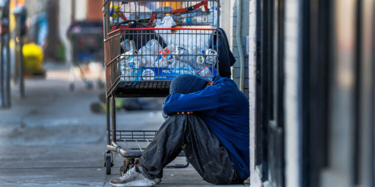 A person in worn clothes sits on a sidewalk with their head on their knees, next to a shopping cart filled with bottles and bags, highlighting the urgent need for access to proper treatment and support.