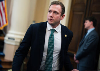 Max Miller wears a dark suit, light dress shirt, and patterned green tie while walking through a government building. An American flag and another man in a suit are visible in the blurred background.