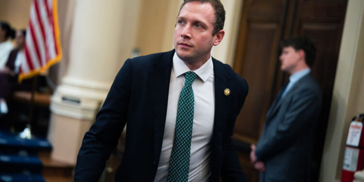 Max Miller wears a dark suit, light dress shirt, and patterned green tie while walking through a government building. An American flag and another man in a suit are visible in the blurred background.