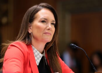 A woman with long brown hair in a red blazer sits at a table with a microphone, appearing to listen attentively during a formal meeting or hearing.