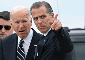 Two men in suits stand outdoors. One is looking forward with his mouth slightly open, while the other gestures and points ahead. A third man in sunglasses stands behind them. A vehicle is visible in the background.