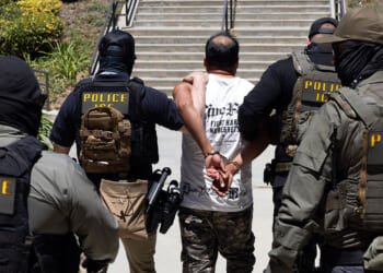 Four officers in POLICE ICE vests escort a handcuffed man up outdoor steps. The officers wear tactical gear and masks, while the man wears a white t-shirt and camouflage pants. The scene appears tense and controlled.