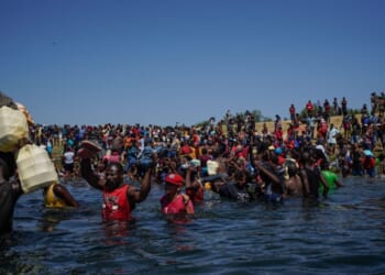 A large crowd of Haitian immigrants wade through waist-deep water, carrying belongings above their heads, while more people gather on the riverbank under a clear blue sky.