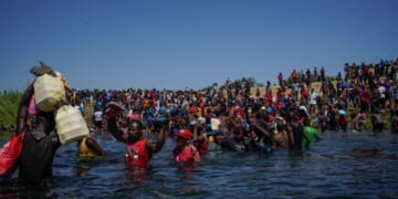 A large crowd of Haitian immigrants wade through waist-deep water, carrying belongings above their heads, while more people gather on the riverbank under a clear blue sky.