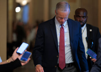 John Thune in a suit and tie looks down while walking indoors, surrounded by people holding smartphones—many of whom appear to be independents covering the event. A security guard walks behind him. The background is softly lit and blurred.
