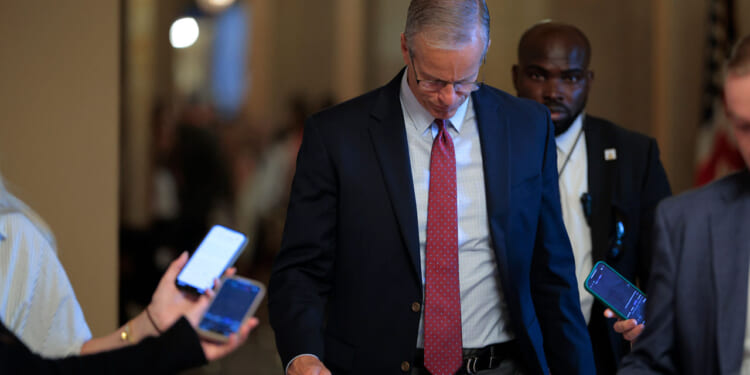 John Thune in a suit and tie looks down while walking indoors, surrounded by people holding smartphones—many of whom appear to be independents covering the event. A security guard walks behind him. The background is softly lit and blurred.