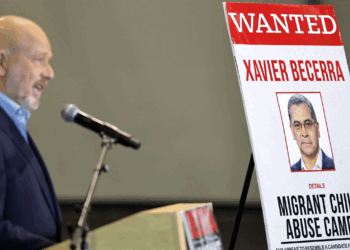A man speaks at a podium next to a large sign reading WANTED with a photo of Xavier Becerra and the words Migrant Child Abuse Camps in bold letters.
