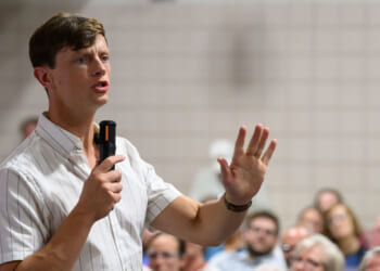 Rob Sand in a striped shirt holds a microphone and speaks, gesturing with his hand, in front of a seated audience in a room with tiled walls.