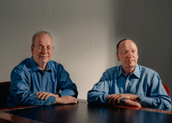 Steven Brill and L. Gordon Crovitz sit at a brown table in front of a gray wall.