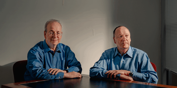 Steven Brill and L. Gordon Crovitz sit at a brown table in front of a gray wall.