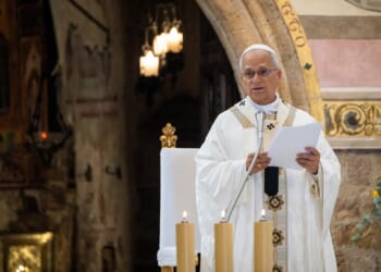 A clergy member in white and gold robes stands at a lectern, holding papers and speaking in a decorated church. Three lit candles and a flower arrangement are visible nearby.