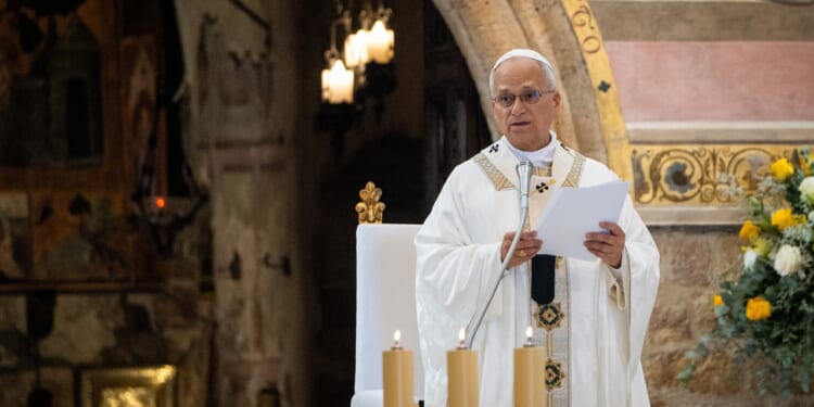 A clergy member in white and gold robes stands at a lectern, holding papers and speaking in a decorated church. Three lit candles and a flower arrangement are visible nearby.