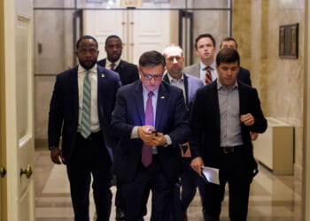 A group of men, including Mike Johnson, in business attire walk down a hallway. The man at the front is looking at his phone, while others follow closely behind. The setting appears to be an official or government building.