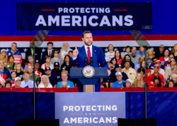Vice President JD Vance in a suit speaks at a podium labeled PROTECTING AMERICANS with a crowd seated behind him and a large sign displaying the same message.