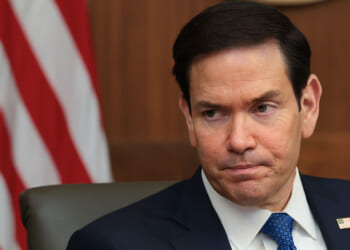 Secretary of State Marco Rubio designated the A/I Collective a terrorist group, noting its support for Jane's Revenge and other leftist terrorism. Pictured: Rubio, a man in a suit and tie sits in front of an American flag, looking to the side with a serious expression. An American flag pin is visible on his lapel.