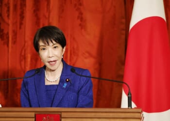 A woman in a blue suit speaks at a podium with microphones, standing beside a Japanese flag and a red curtain backdrop.