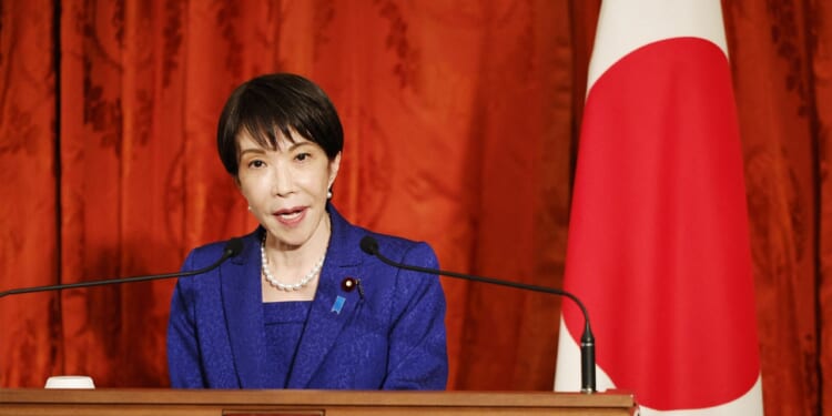 A woman in a blue suit speaks at a podium with microphones, standing beside a Japanese flag and a red curtain backdrop.