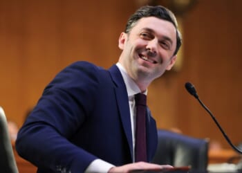 Senator Jon Ossoff appears in a navy suit and red tie. He smiles while seated at a desk with a microphone, in a formal wood-paneled room.