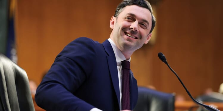 Senator Jon Ossoff appears in a navy suit and red tie. He smiles while seated at a desk with a microphone, in a formal wood-paneled room.