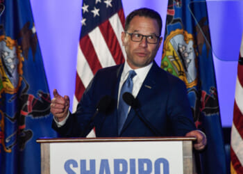 A man in a blue suit, identified as Josh Shapiro, speaks at a podium labeled SHAPIRO, with American and Pennsylvania state flags in the background. He gestures with one hand and looks to the side.