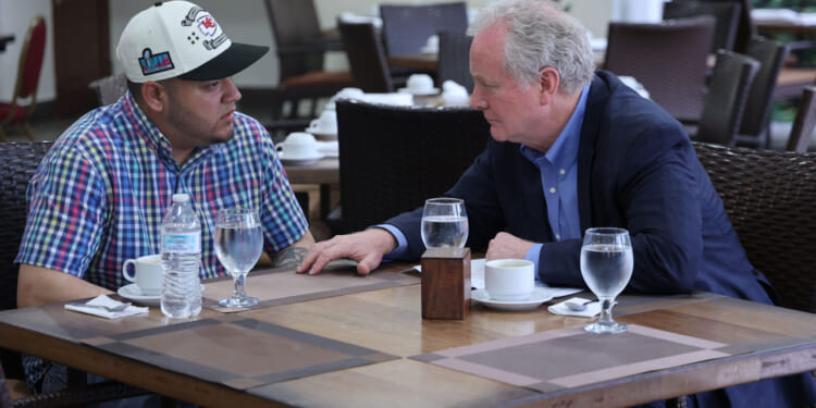 Kilmar Abrego Garcia and Sen. Chris Van Hollen sit at a restaurant table having a serious conversation. Garcia in a plaid shirt and cap, faces Van Hollen, who is dressed in a suit. Water glasses, cups, and a water bottle are on the table. The background shows empty chairs and tables.