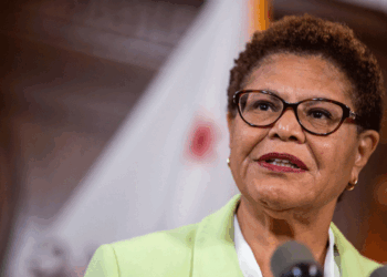 A woman with short hair and glasses speaks at a podium. She is wearing a light green blazer, and there is a blurred bass guitar resting in the background alongside the flag.