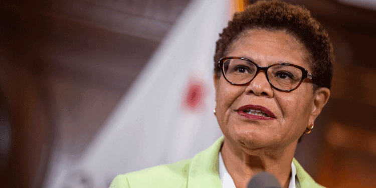 A woman with short hair and glasses speaks at a podium. She is wearing a light green blazer, and there is a blurred bass guitar resting in the background alongside the flag.