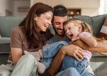 A family is pictured with a mother, father, and daughter laughing in their living room.