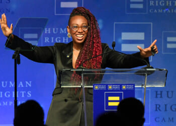 A person with long braids, glasses, and a black blazer stands smiling with arms raised at a podium, speaking at a Human Rights Campaign event. Blue backdrop with campaign logos is visible.