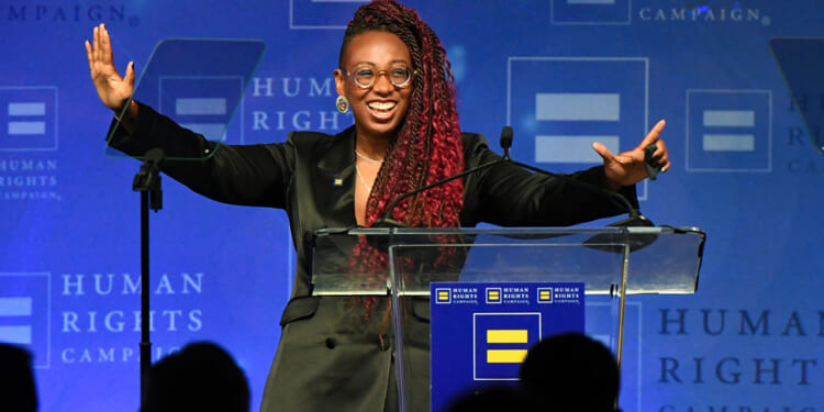 A person with long braids, glasses, and a black blazer stands smiling with arms raised at a podium, speaking at a Human Rights Campaign event. Blue backdrop with campaign logos is visible.