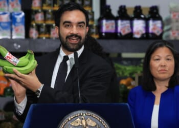New York City Mayor Zohran Mamdani in a suit at a podium holds up a bunch of green bananas, while a woman in blue stands beside him. Shelves with groceries are visible in the background.
