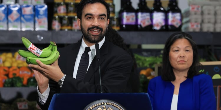 New York City Mayor Zohran Mamdani in a suit at a podium holds up a bunch of green bananas, while a woman in blue stands beside him. Shelves with groceries are visible in the background.